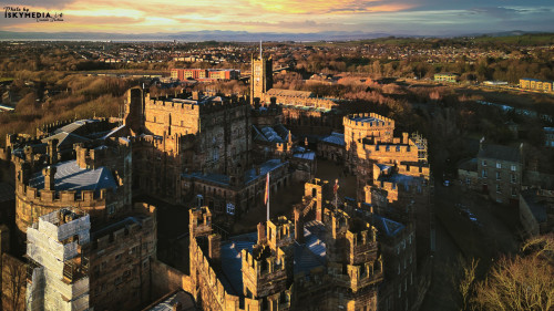 Aerial view of a historic castle at sunset with surrounding landscape and town under a golden sky in Lancaster.