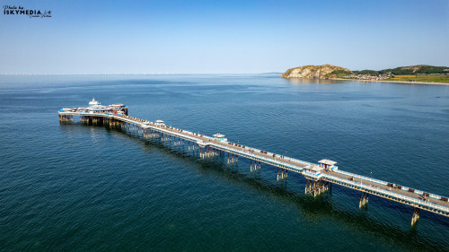 Aerial shot of Llandudno Pier extending into the sea, with a clear blue sky and distant hills in the background.