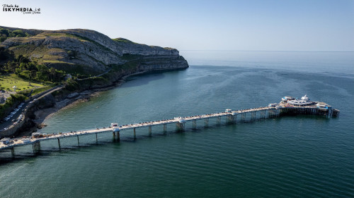 Aerial view of Llandudno Pier extending into the sea, with cliffs and greenery in the background.