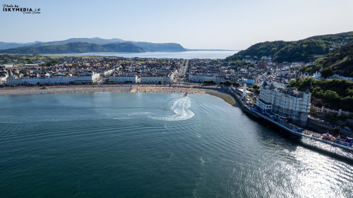 Aerial view of a picturesque coastal town with a sandy beach, waterfront buildings, and a body of water reflecting sunlight in Llandudno, North Wales.