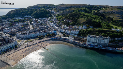 An aerial view of a coastal town with a sandy beach, a pier, and cliffs in the background, showcasing a vibrant seaside setting in Llandudno, North Wales.
