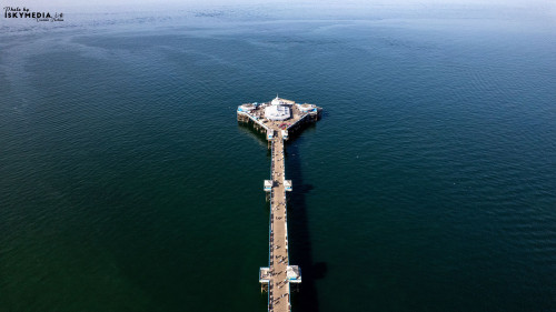 An aerial view of a pier extending into a vast, deep blue ocean, with a clear sky above in Llandudno, North Wales.