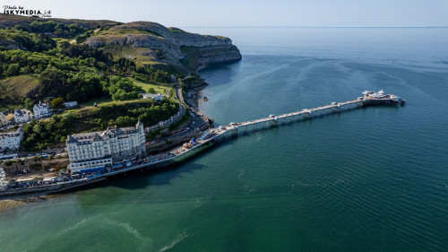 Aerial view of a coastal town with a pier extending into the sea, surrounded by cliffs and lush greenery in Llandudno, North Wales.