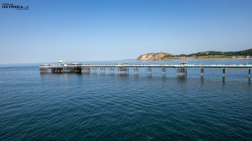 A scenic view of Llandudno Pier extending into the sea, with a backdrop of coastal hills and a clear blue sky.