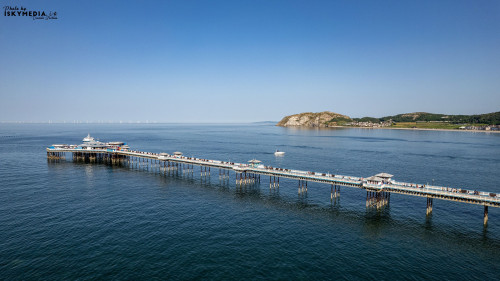 A scenic view of Llandudno Pier extending into the sea, with a backdrop of coastal hills and a clear blue sky.